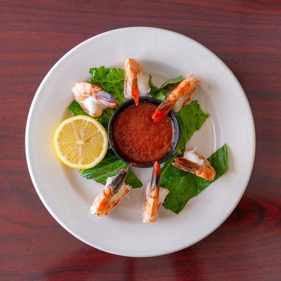Cooked shrimp arranged around cocktail sauce, lettuce leaves, and a lemon half on a white plate.