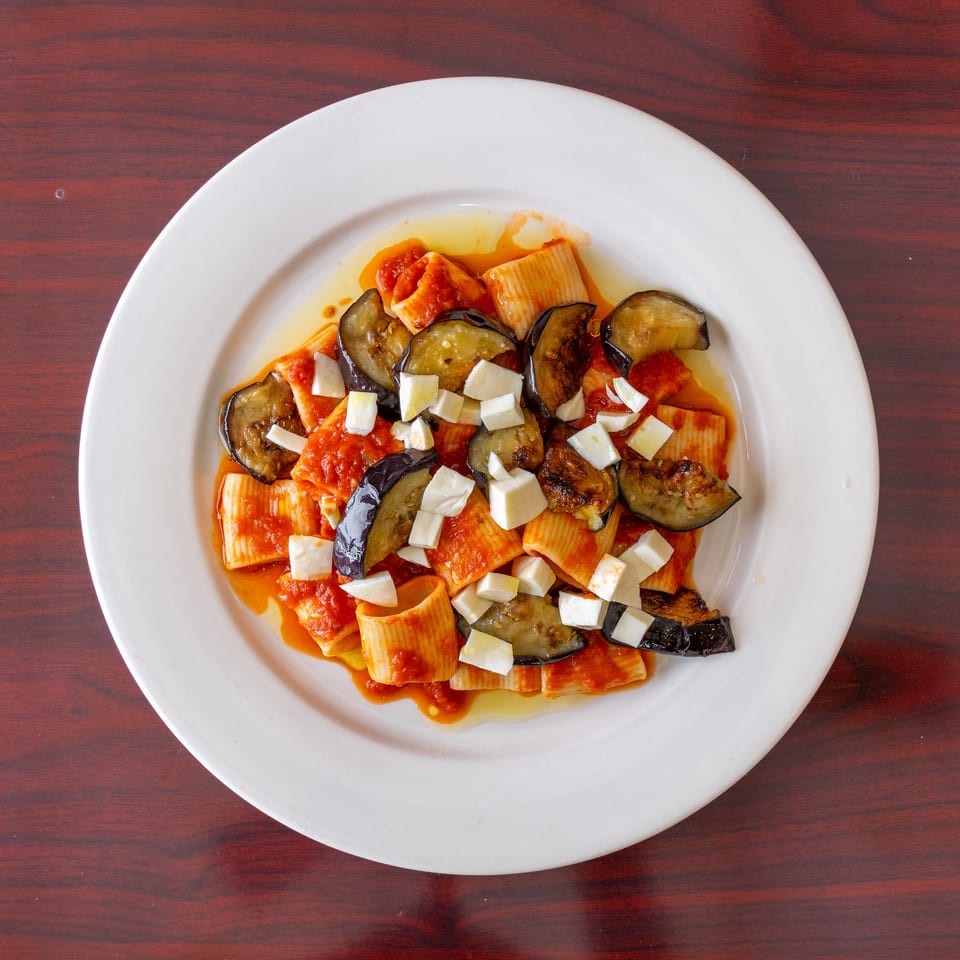 Rigatoni pasta with tomato sauce, roasted eggplant, and diced white cheese on a white plate.