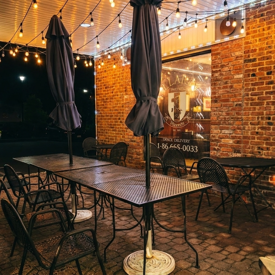 Outdoor restaurant patio at night with brick walls, black tables and chairs, closed umbrellas, and glowing string lights.