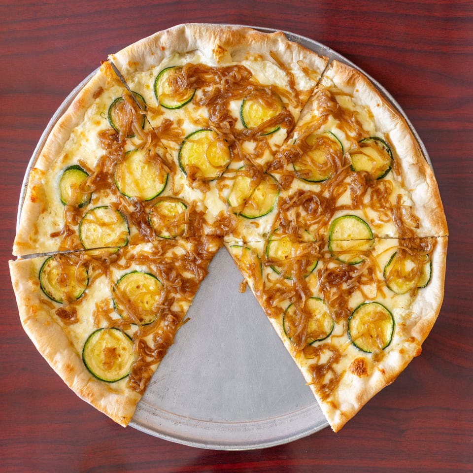 Thin-crust pizza topped with melted cheese, zucchini slices, and caramelized onions on a metal tray.