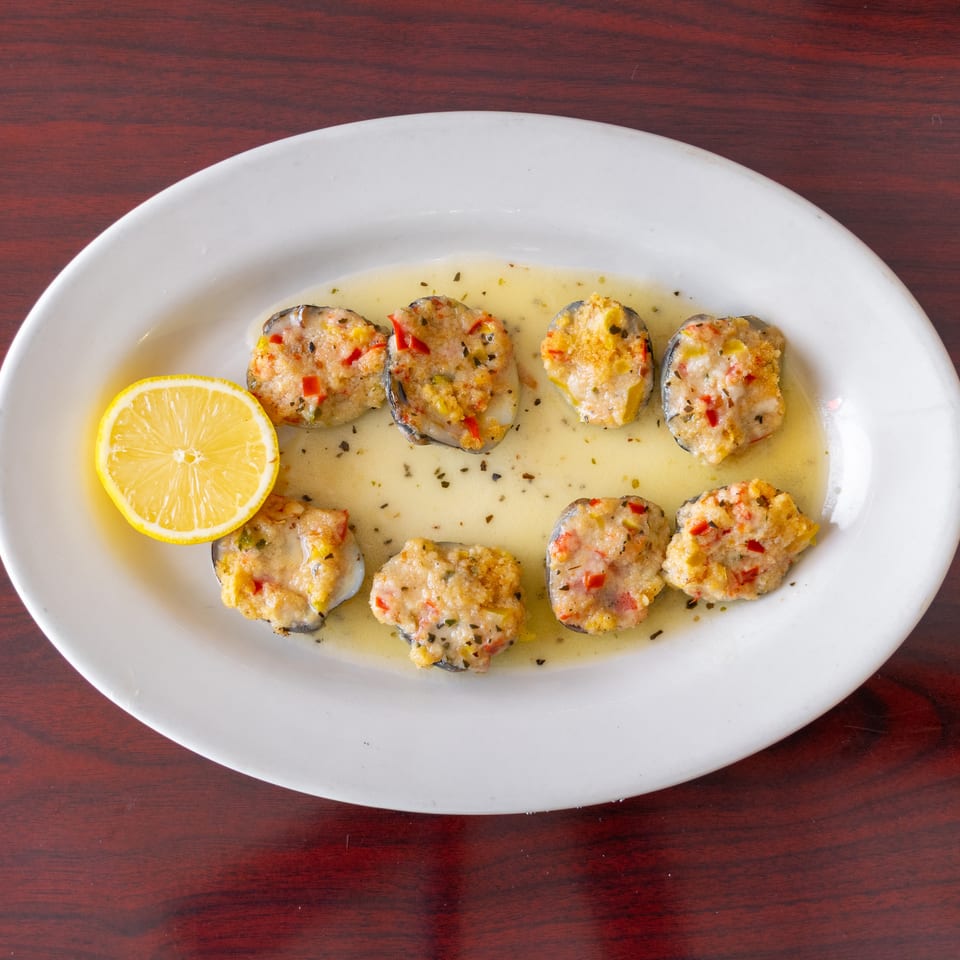 Baked stuffed mussels with peppers in lemon butter sauce, garnished with a lemon half on a white platter.