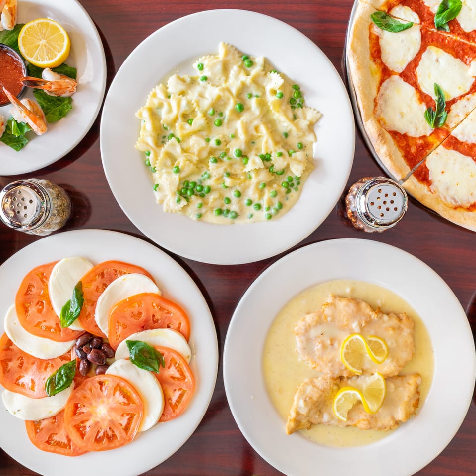 Overhead spread of Italian dishes including shrimp, creamy bow-tie pasta with peas, margherita pizza, caprese salad, and lemon chicken.