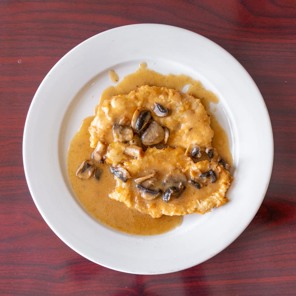 Breaded chicken cutlets topped with mushrooms and brown gravy on a white plate over a wooden table.