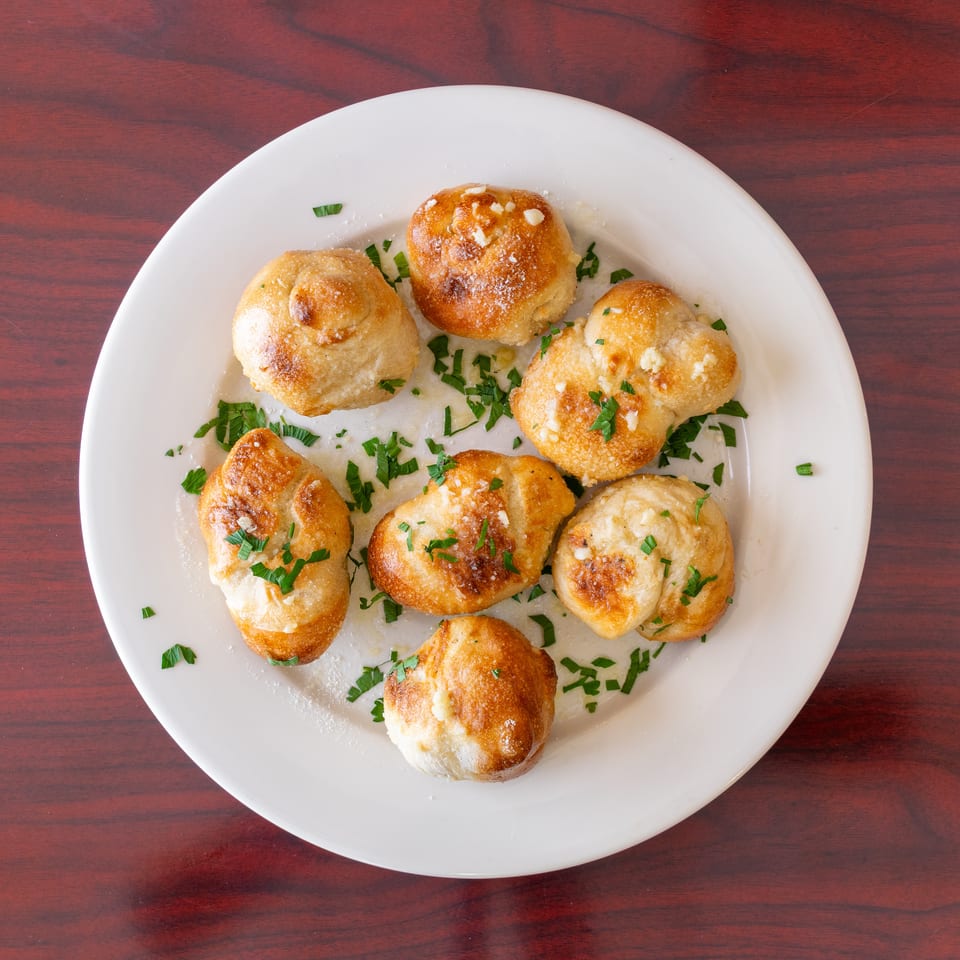 Golden-brown garlic rolls garnished with chopped parsley on a white plate atop a dark wooden table.