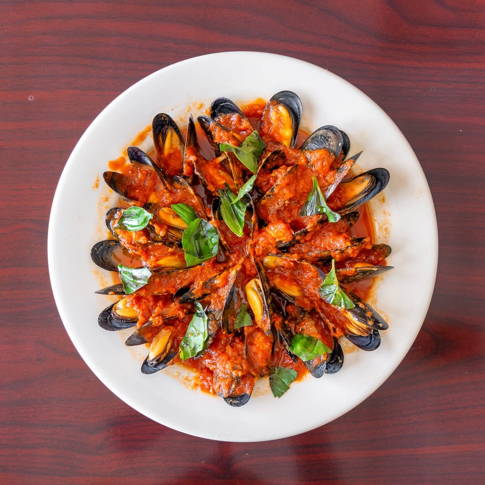 Open mussels coated in tomato sauce and garnished with fresh basil on a white plate atop a wooden table.