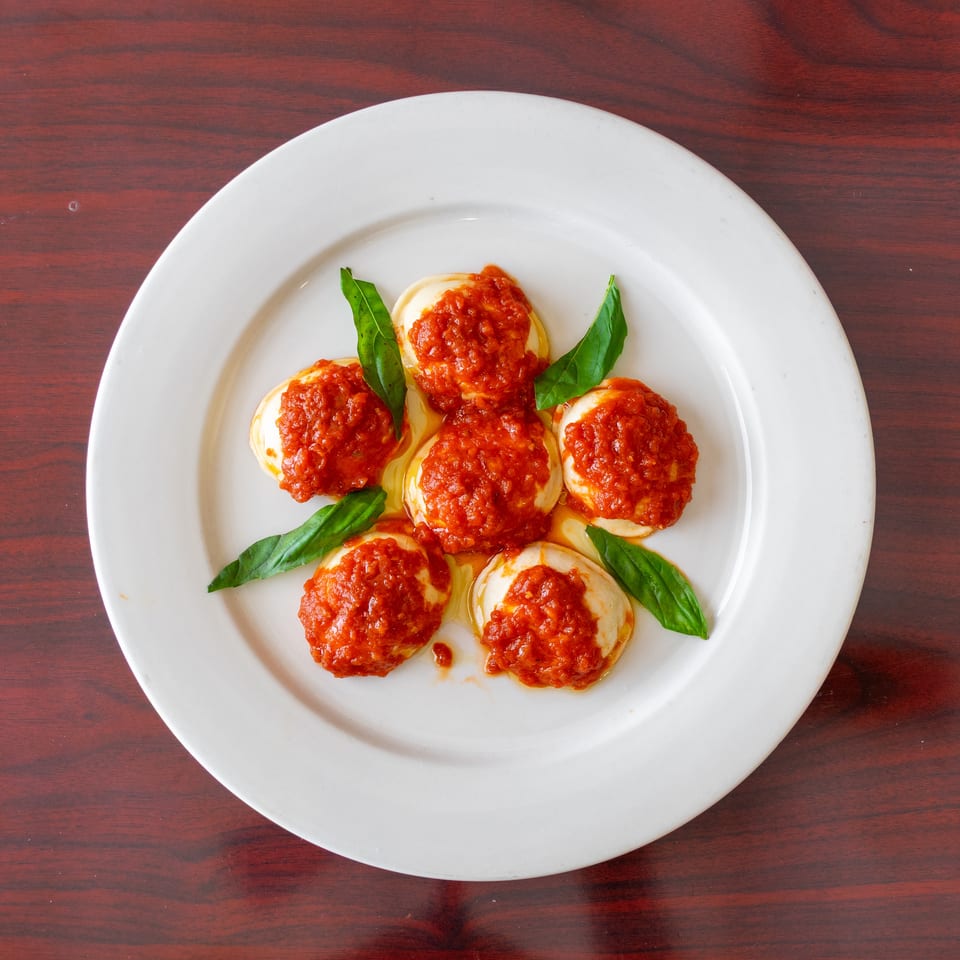 Five round pasta pieces topped with chunky tomato sauce and basil leaves on a white plate over a wooden table.