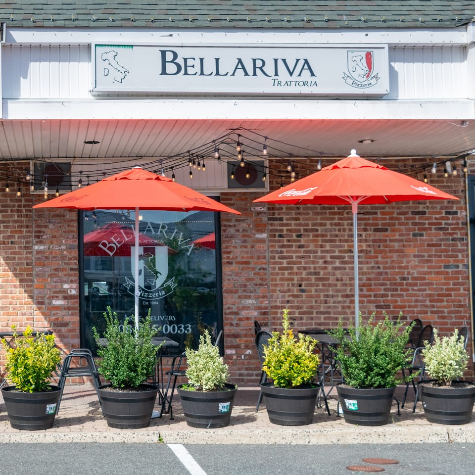 Bellaria Trattoria pizzeria storefront with red umbrellas, string lights, brick walls, outdoor seating, and potted shrubs.