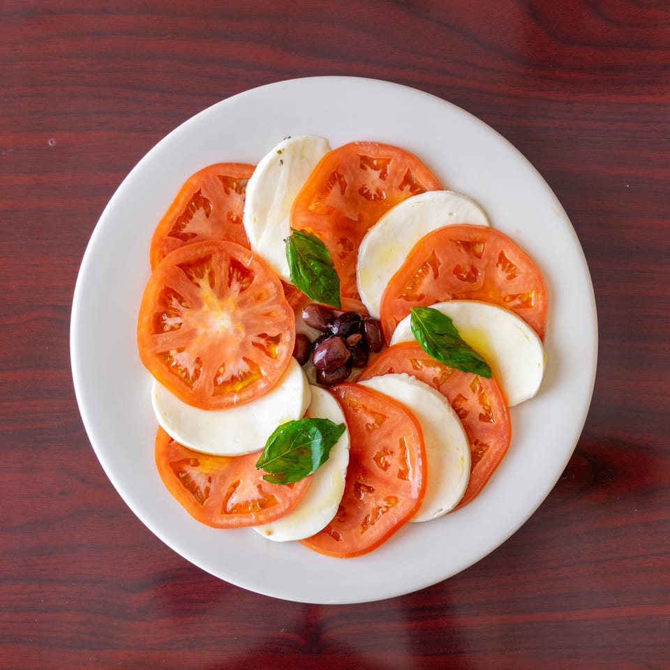 Caprese salad with sliced tomatoes, fresh mozzarella, basil leaves, and olives on a white plate.